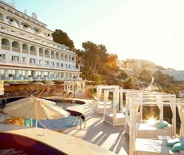 Hotellfasad i vit med arkader, swimmingpool och solstolar med parasoller och dagsängar, omgiven av träd och byggnader. (AI-genererad text)