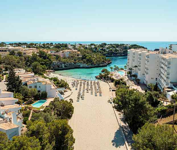 En strand med parasoller omgivet av byggnader och vegetation, med havet i bakgrunden. (AI-genererad text)