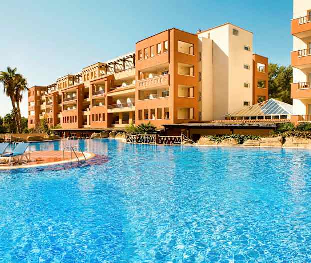 Bilden visar en byggnad vid en stor utomhuspool med blått vatten och solstolar placerade vid poolkanten. I bakgrunden syns några palmer. (AI-genererad text)