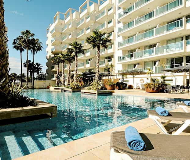 Bild av en hotellpool med solstolar och palmer omgivna av ett modern byggnadskomplex. (AI-genererad text)