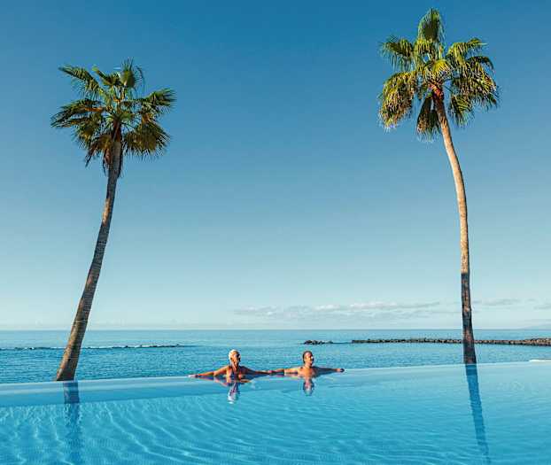 Två personer i en infinitypool med utsikt över havet och två palmer. (AI-genererad text)