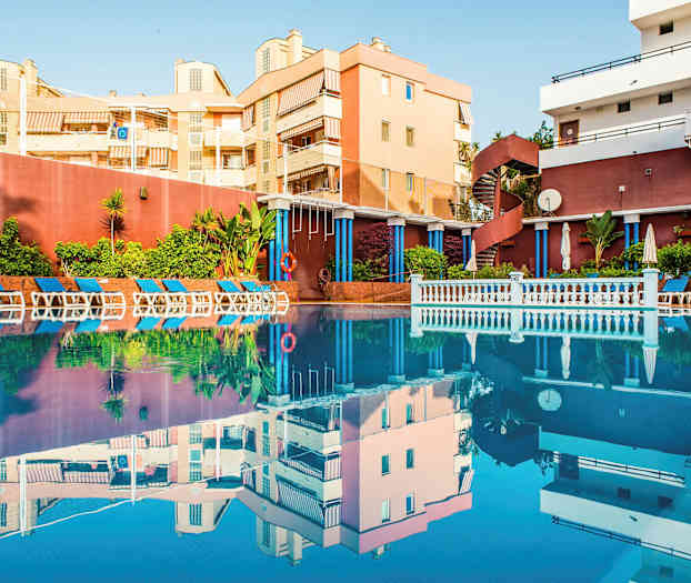 En utomhuspool omgiven av solstolar och gröna växter, med byggnader i bakgrunden. (AI-genererad text)