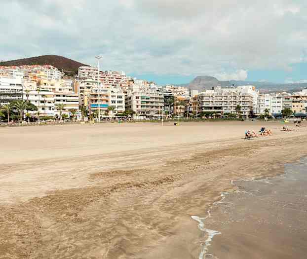 Strand med sand och vågor, omgiven av byggnader och kullar i bakgrunden. (AI-genererad text)