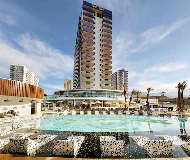 Ett hotell med en stor pool och omgivande byggnader under en blå himmel. (AI-genererad text)