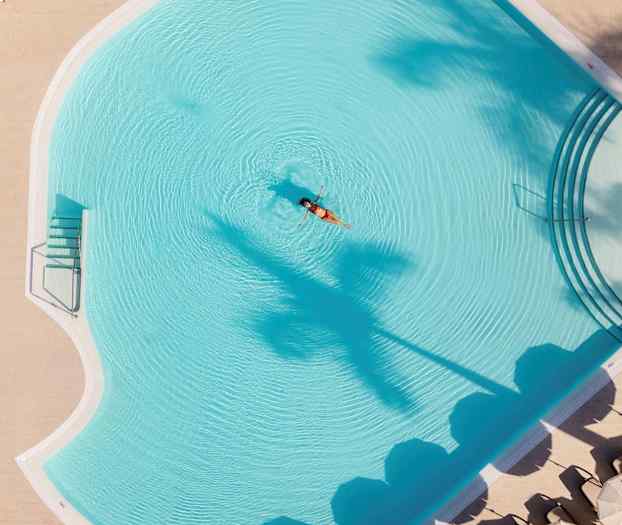 En kvinna flyter i en hotellpool, omgiven av parasoller och palmer. (AI-genererad text)