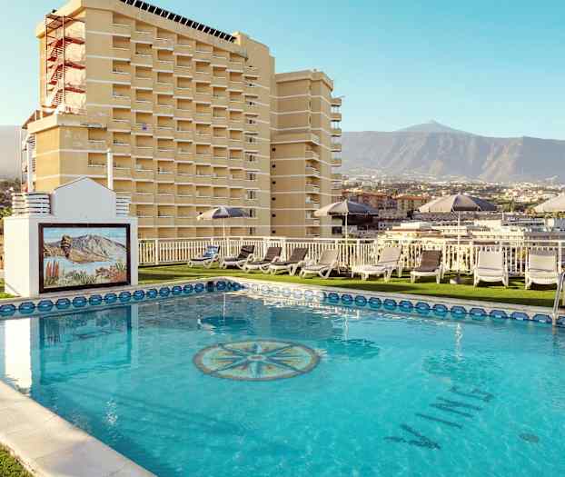 En hotellpool med solstolar och utsikt mot berg och byggnader. (AI-genererad text)