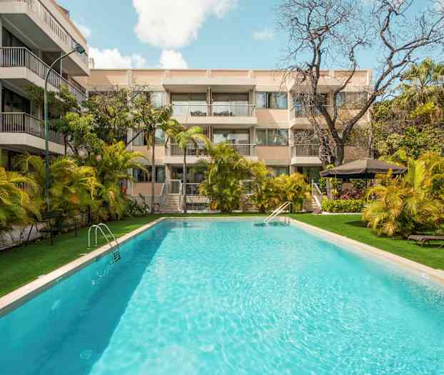 En utomhuspool som är omgiven av palmer och en byggnad med balkonger. (AI-genererad text)