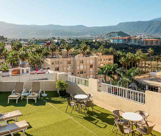 Bild av en hotellets takterrass med solstolar och bord, omgiven av gröna områden och palmer, med utsikt över omgivande stadsområde och berg. (AI-genererad text)