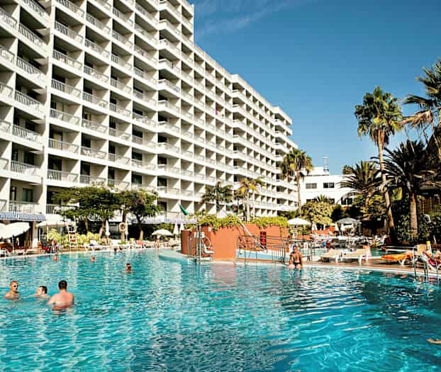 Stor hotellbyggnad med många balkonger, omgiven av en pool och palmer. (AI-genererad text)