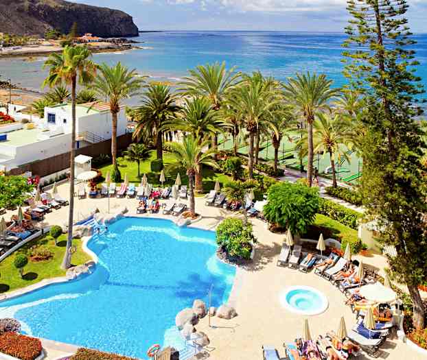 Hotellpool med solsängar och parasoller, omgiven av tropiska palmer och kustlandskap. (AI-genererad text)