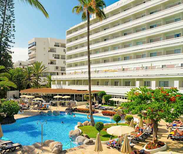 Ett hotell med en pool i förgrunden, omgiven av solstolar och parasoller samt grönska. (AI-genererad text)