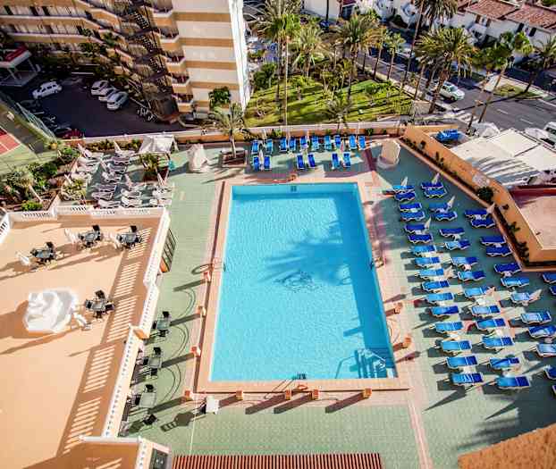 Vy över en pool och solstolar vid ett hotell, med utsikt över omgivande byggnader och grönska. (AI-genererad text)