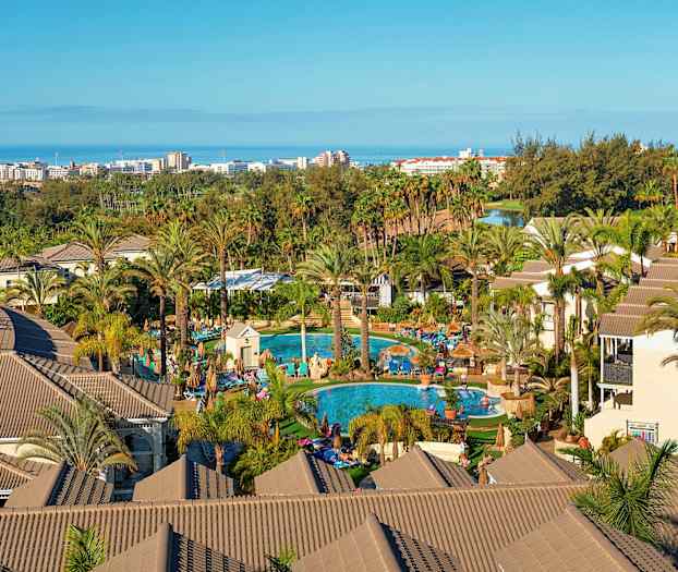 En vy över ett semesterhotell med palmer, pooler och byggnader, i bakgrunden syns havet och en stad. (AI-genererad text)