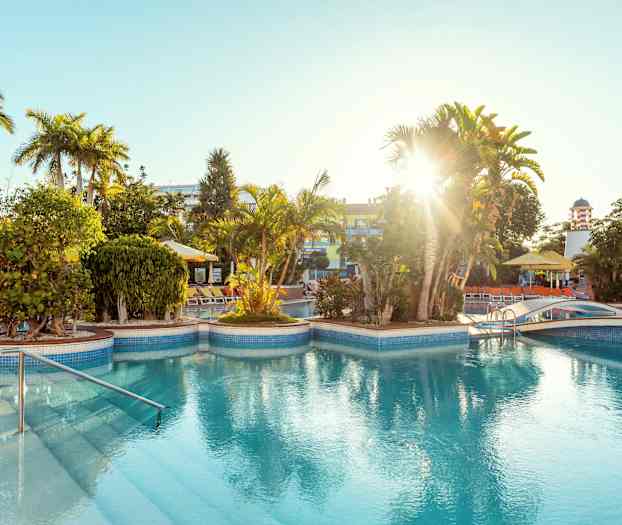 En utomhuspool med omgivande solstolar, gröna växter och palmer under starkt solljus. (AI-genererad text)