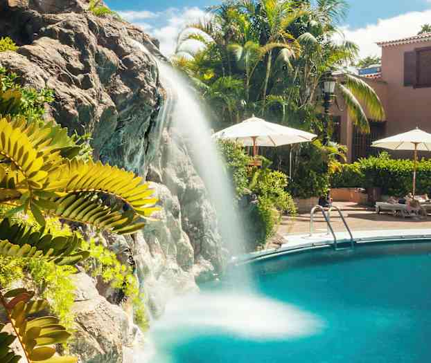 Vattenfall som rinner ner i en swimmingpool, med omgivande grönska och solstolar. (AI-genererad text)