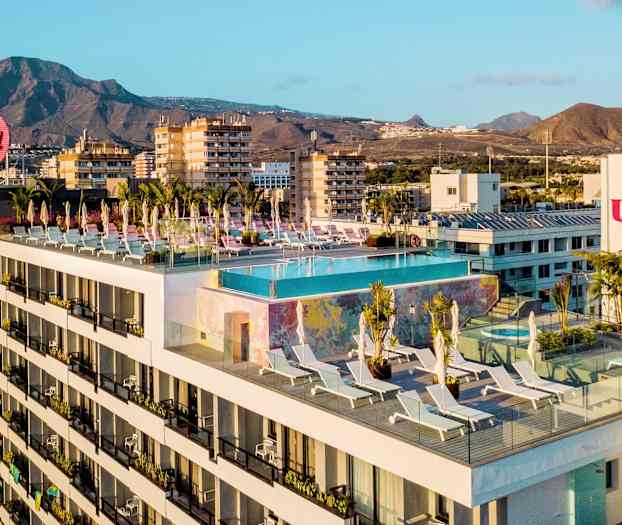 Hotellbyggnad med en infinitypool och solstolar på takterrassen, i bakgrunden berg och städer. (AI-genererad text)