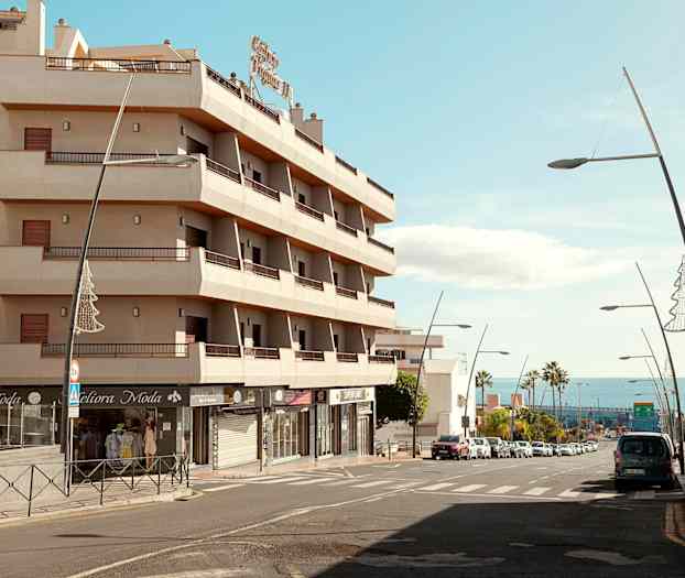 En byggnad med balkonger vid en gata med bilar och gatlampor, med havet i bakgrunden. (AI-genererad text)