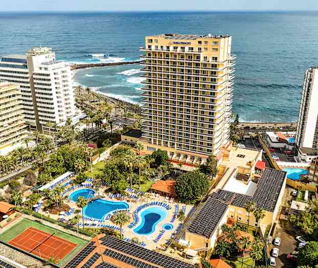 Hotellanläggning med poolområde och tennisbana, vid havet, omgiven av höga byggnader. (AI-genererad text)