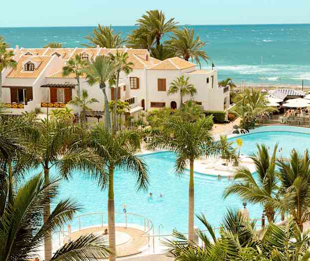 Hotellbyggnader med terrakottafärgade tak vid en pool omgiven av palmer och hav i bakgrunden. (AI-genererad text)