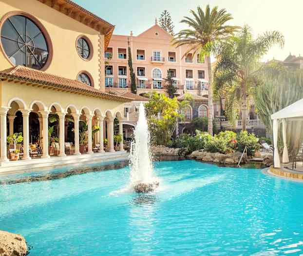 Hotell byggnad med en pool och fontän i förgrunden, omgiven av palmer och grönska. (AI-genererad text)