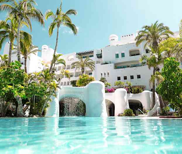 Vit byggnad med palmer omkring och pool i förgrunden. (AI-genererad text)