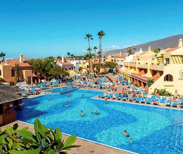 Stor utomhuspool omgiven av solstolar och byggnader med berg i bakgrunden. (AI-genererad text)