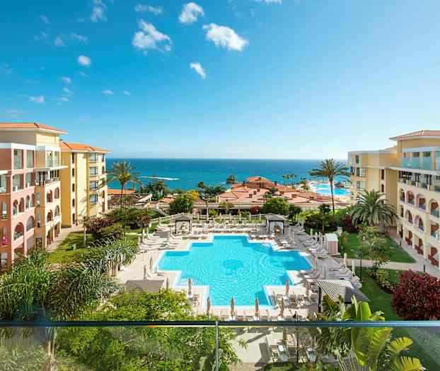 Bild på ett hotellområde med en pool, omgiven av solstolar och parasoller. Hotellets byggnader är placerade på båda sidor och i bakgrunden syns havet. (AI-genererad text)