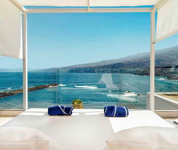 En utsikt från en terrass med liggbäddar, glasskydd, två blå handdukar och ett martiniglas i förgrunden med hav, klippor och berg i bakgrunden. (AI-genererad text)