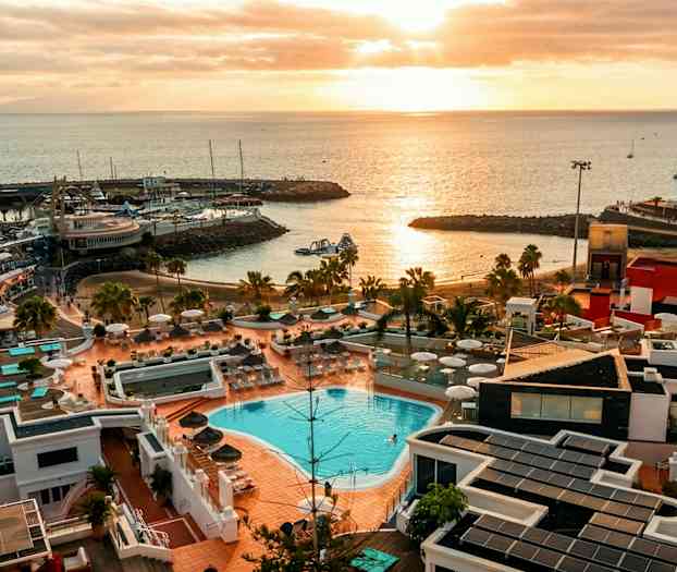 Utsikt över ett kustområde vid solnedgången med en anläggning som har pool och solterrass i förgrunden samt havet och småbåtar i bakgrunden. (AI-genererad text)