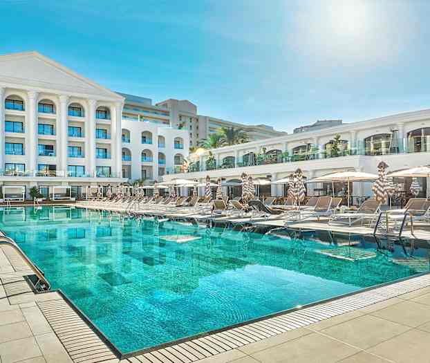 En hotellbyggnad med en stor swimmingpool och solstolar framför. (AI-genererad text)