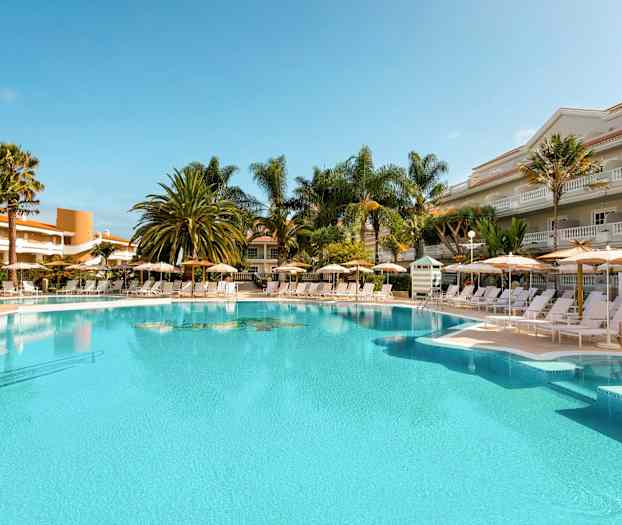 Utomhuspool med solstolar och parasoller omgivna av palmer och byggnader. (AI-genererad text)