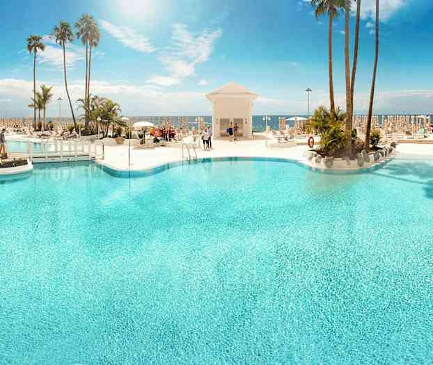 En stor utomhuspool omgivet av solstolar och palmer med havet i bakgrunden. (AI-genererad text)