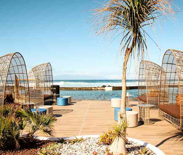 Utemöbler med rottingdesign står på en träplattform vid havet. (AI-genererad text)