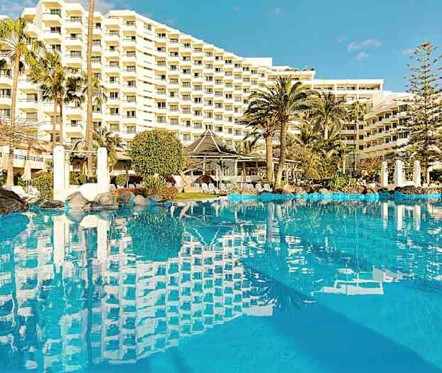 En bild av en utomhuspool omgiven av palmer, samt ett stort hotell i bakgrunden. (AI-genererad text)