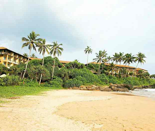 En sandstrand med grönt buskage och palmer i bakgrunden. Till höger syns en byggnad med gula väggar. (AI-genererad text)