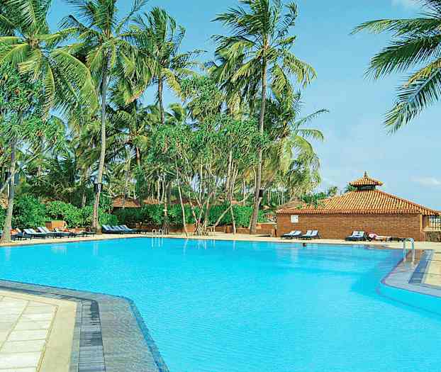 En stor pool omgiven av höga palmer och solstolar nära en byggnad med tegeltak. (AI-genererad text)