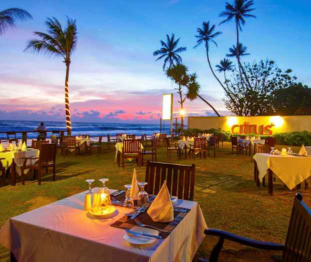 En restaurang utomhus med vita dukar och trästolar vid solnedgången nära havet. (AI-genererad text)