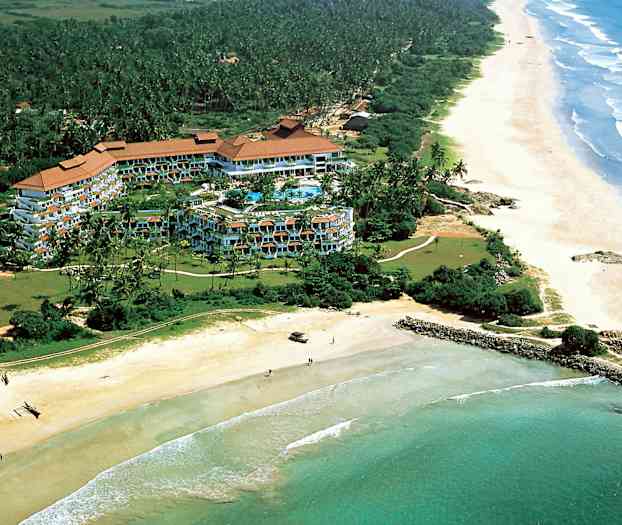 Flygbild av ett hotellkomplex omgivet av grönska vid kusten med en lång sandstrand och hav. (AI-genererad text)