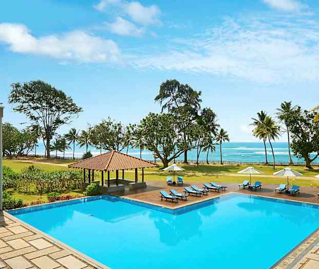 En utomhuspool med solstolar och parasoller, omgiven av en gräsmatta och träd, med utsikt över stranden och havet. (AI-genererad text)