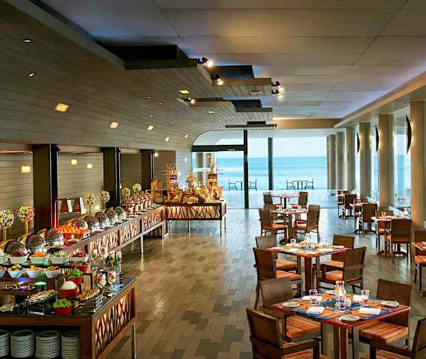 Bild av en restaurang med dukade bord och en buffé fylld med olika maträtter, samt stora fönster som vetter ut mot havet. (AI-genererad text)