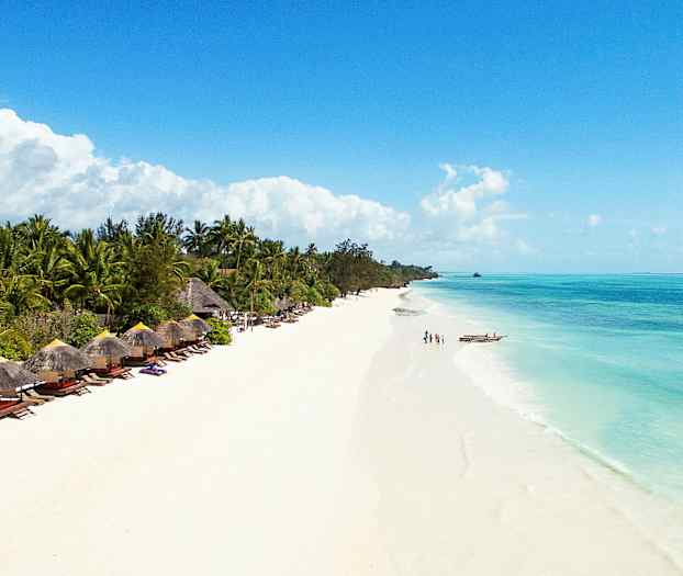 En strand med vit sand och klart blått vatten, omgiven av grönskande palmer och parasollstolar. (AI-genererad text)