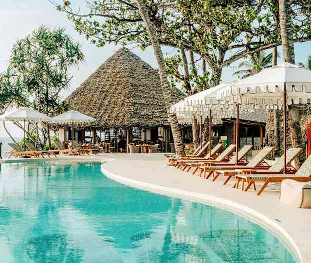 En poolområde med solstolar och parasoller. I bakgrunden syns en byggnad med ett halmtak och palmer. (AI-genererad text)