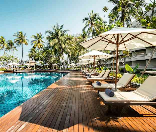 En pool med klart vatten omgivet av solstolar och parasoller på ett trädeck. Palmer finns i bakgrunden. (AI-genererad text)