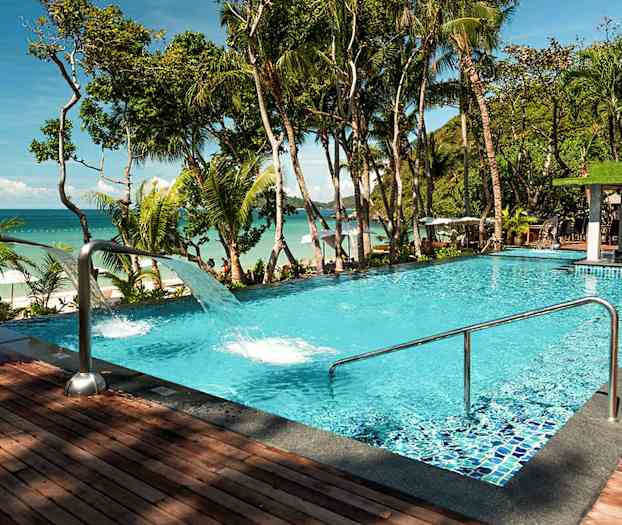 En pool med klart blått vatten, omgiven av tropiska träd och med utsikt över havet. (AI-genererad text)