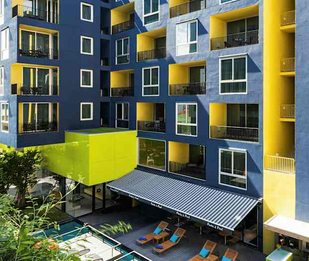 Byggnad med blå och gula fasader samt balkonger, framför en pool och solstolar. (AI-genererad text)