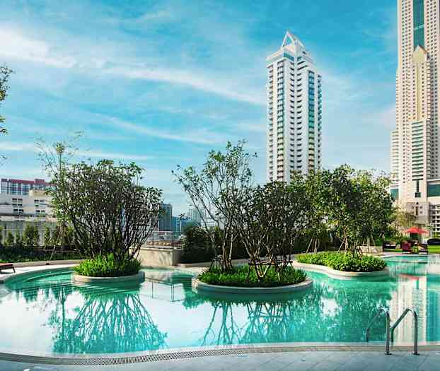 En pool med klarblått vatten omgiven av grönska och höga byggnader i bakgrunden. (AI-genererad text)