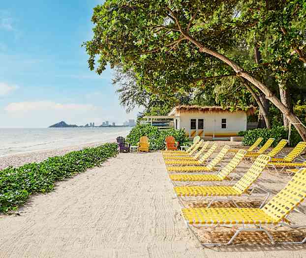Strand med solstolar och grönska samt hus i bakgrunden. (AI-genererad text)