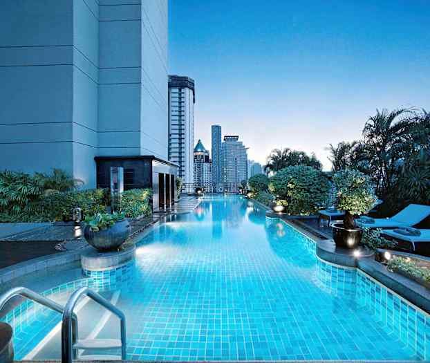 En utomhuspool vid en byggnad omgiven av grönska, med solstolar och höga byggnader i bakgrunden. (AI-genererad text)