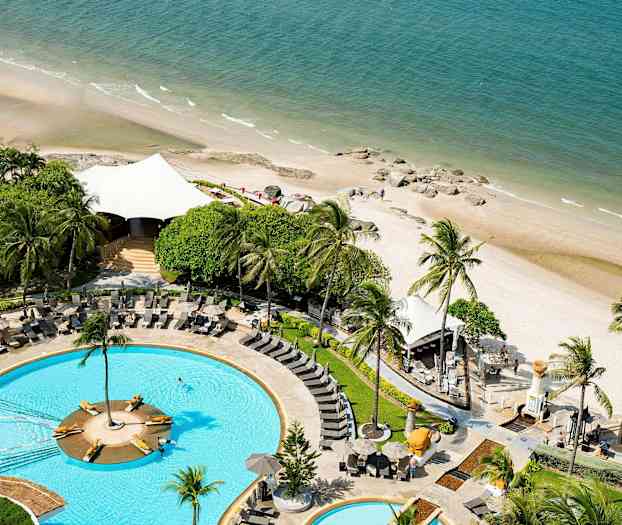 Bild av en pool med solstolar omgiven av grönska, belägen bredvid en sandstrand som möter havet. (AI-genererad text)