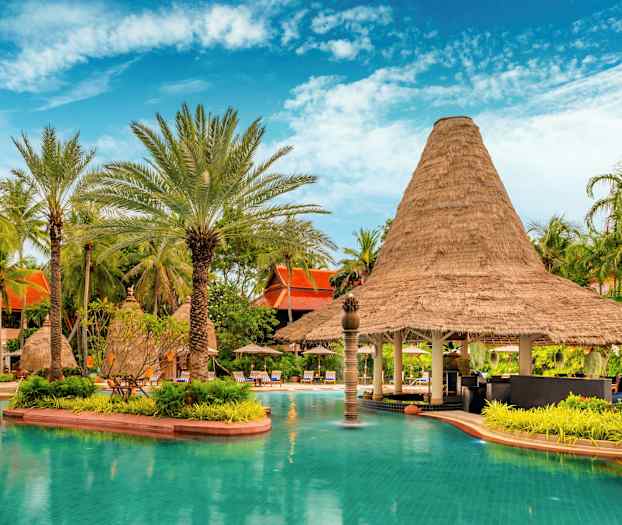 En pool med turkost vatten omgiven av grönskande palmer och en byggnad med halmtak. (AI-genererad text)